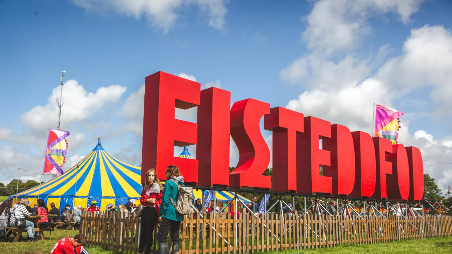Gŵyl ddiwylliannol yr Eisteddfod yng Nghymru, gyda’r mynychwyr yn y llun o flaen arddangosfa wedi’i llunio â thestun a phabell yr ŵyl a baneri y tu ôl iddynt.
